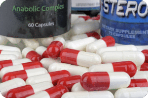 Cápsulas ou pílulas de suplementos nas cores branco e vermelho, espalhadas sobre a mesa, com potes de suplementos com rótulos indicando "Anabolic Complex" e "Steroid" ao fundo. A imagem simboliza nutracêuticos, suplementação e suporte anabólico.