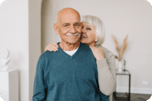 Casal idoso feliz em um ambiente interno. O homem, careca e com bigode, sorri para a câmera vestindo um suéter azul. A mulher, com cabelos curtos e grisalhos, o abraça por trás e sorri, transmitindo afeto e bem-estar na longevidade.