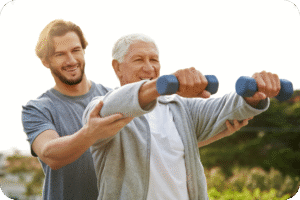 Homem idoso sorridente, com cabelo branco, levantando halteres leves sob a supervisão e assistência de um personal trainer mais jovem, com barba. Ambos estão ao ar livre, em um dia ensolarado, representando o exercício físico na terceira idade e a importância do treinamento de força para a longevidade saudável.