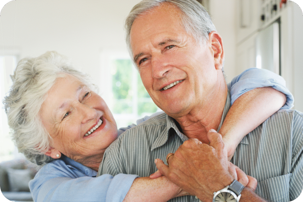 Casal idoso feliz e sorridente, abraçado em um ambiente doméstico claro. A mulher, com cabelos brancos, abraça o homem por trás, que usa um relógio de pulso. A imagem representa o afeto e a qualidade de vida na Longevidade Saudável.