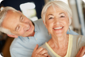 Casal idoso feliz, com a mulher sorrindo para a câmera em primeiro plano e o homem, também sorrindo, gentilmente apoiado nela por trás. Ambos têm cabelos grisalhos e transmitem uma sensação de bem-estar e companheirismo.