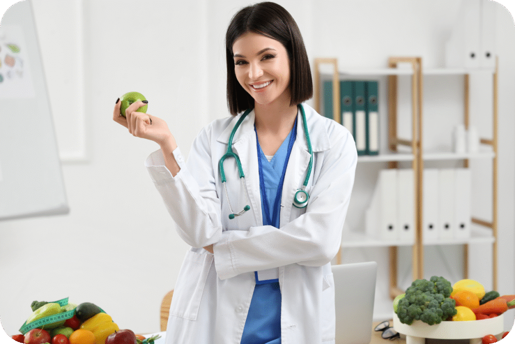 Médica nutróloga (ou nutricionista) sorrindo, vestindo jaleco branco e estetoscópio, segurando uma maçã verde no consultório, com frutas e vegetais sobre a mesa.