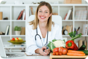 Mulher, com aparência de médica ou nutricionista, sorrindo e olhando para frente em um consultório ou escritório. Ela veste um jaleco branco e estetoscópio. À sua frente, há um laptop aberto e uma tábua de madeira com diversos vegetais frescos como tomate, pimentão, cenoura e couve-flor.