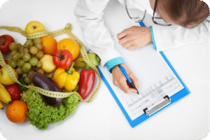 Vista superior de um profissional de saúde (médico ou nutricionista) vestindo jaleco, escrevendo com uma caneta azul em uma prancheta com um plano de dieta ou prontuário. À esquerda, há um monte de frutas e vegetais coloridos, como uvas, laranjas, pimentões e alface, com uma fita métrica amarela envolvendo-os.