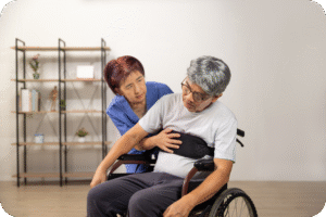 Pessoa idosa sentada em uma cadeira de rodas, sendo auxiliada por um cuidador ou profissional de saúde. A imagem ilustra a perda de mobilidade e a dependência causada pela sarcopenia (perda de massa muscular).