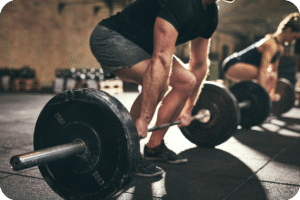 Duas pessoas, um homem e uma mulher, realizando levantamento de peso (deadlift ou agachamento com barra) em uma academia. A imagem foca no treino de força e na sobrecarga progressiva para ganho muscular após os 40.