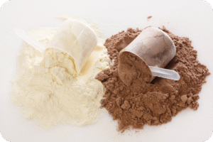 Duas porções de pó de suplemento, uma branca (como baunilha ou sem sabor) e outra marrom (como chocolate), cada uma com uma colher-medida plástica inserida. A imagem destaca a suplementação proteica, relevante para a otimização muscular.